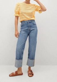 Top de ganchillo amarillo con manga abullonada, combinado con unos vaqueros de tiro alto azules con dobladillos recortados y sandalias de dedo marrones.
