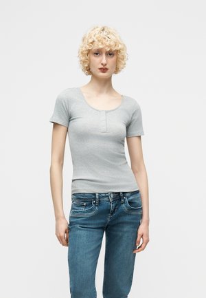 Νεαρό άτομο με κοντά σγουρά ξανθά μαλλιά, που φοράει γκρι κοντομάνικη μπλούζα henley και μπλε τζιν, στέκεται μπροστά σε απλό φόντο.