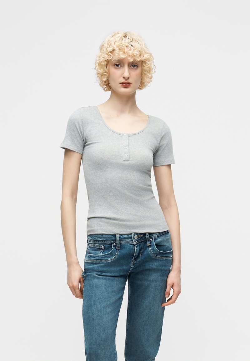 Νεαρό άτομο με κοντά σγουρά ξανθά μαλλιά, που φοράει γκρι κοντομάνικη μπλούζα henley και μπλε τζιν, στέκεται μπροστά σε απλό φόντο.