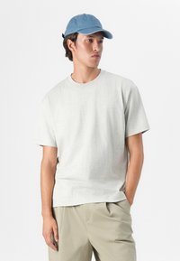 Hellgraues Baumwoll-T-Shirt mit Rundhalsausschnitt und kurzen Ärmeln, kombiniert mit hellbeigen Shorts. Der Träger trägt eine blaue Denim-Mütze.