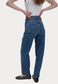 Jeans in denim blu a vita alta con taglio dritto, caratterizzati da due tasche posteriori e una texture scolorita. Indossati con ballerine nere con borchie.