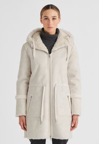 Frau in einem beigen, mit Kapuze versehenen Schafsfellmantel über einem dunkelgrauen Rollkragenpullover, die vor einem schlichten hellen Hintergrund steht.