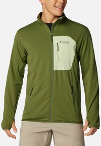 Giacca in pile verde con un design testurizzato, tasche con zip e un pannello anteriore di colore verde chiaro. Presenta un colletto alto e maniche aderenti.