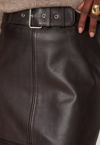 Jupe en cuir marron foncé avec couture verticale et ceinture assortie, portée avec un haut en tricot marron clair et une main montrant une bague en or sur deux doigts.