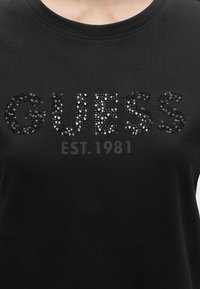 Μαύρο μπλουζάκι με τη λέξη "GUESS" γραμμένη με μαύρες και λευκές στρας και την ένδειξη "EST. 1981" τυπωμένη παρακάτω σε σκούρο γκρι.