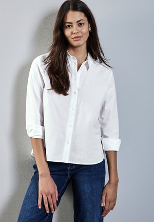 Camicia bianca a bottone con maniche lunghe arrotolate fino ai gomiti, realizzata in tessuto di cotone. Indossata con jeans blu scuro su uno sfondo neutro.