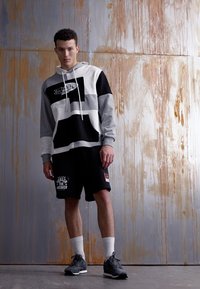 Hnedá, biela a čierna mikina s kapucňou a logom, v kombinácii s čiernymi šortkami s grafickým textom. Model má obuté biele ponožky a tmavé tenisky.