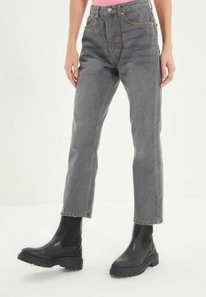 Osoba ubrana w szare jeansy o prostych nogawkach i czarne botki, stojąca na białym tle.