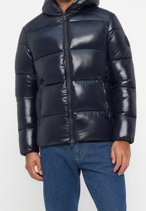 Homme portant une veste matelassée noire brillante avec capuche et fermeture éclair, associée à un jean bleu, debout devant un fond clair uni.