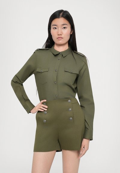 Olive zelena vojaška srajca s sprednjim gumbom in ramenskimi epauletami, v kombinaciji z hlačami visokega pasu, ki imajo dvocertnasto gumbno podrobnost.