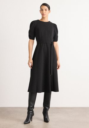 Robe noire avec des manches bouffantes, corsage ajusté et détail de taille nouée. Jupe fluide en A. Portée avec des bottes noires mi-mollet. Texture lisse.
