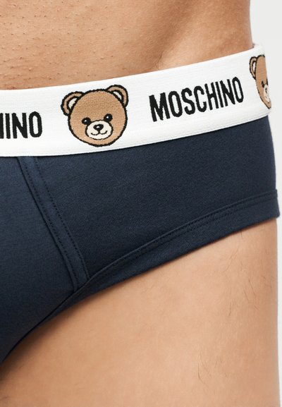 Σλιπ ναυτικού χρώματος με λευκή ζώνη μέσης που φέρει επαναλαμβανόμενο λογότυπο Moschino και χαμογελαστά πρόσωπα αρκουδάκια, φορεμένα στη μέση και το ισχίο ενός ατόμου.