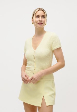 Mujer con cabello rubio, vistiendo un cárdigan corto de manga corta y textura suave de color amarillo pálido, y una minifalda a juego con una abertura frontal, sonriendo y mirando hacia un lado.