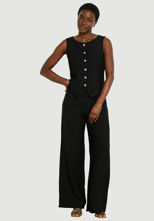 Femme portant un top noir sans manches boutonné et un pantalon noir à jambes larges, debout avec une main sur la hanche sur fond neutre.
