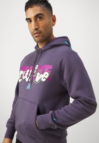 Felpa viola realizzata in misto cotone, con tasca frontale, cappuccio regolabile e un design grafico colorato con testo in rosa e verde.