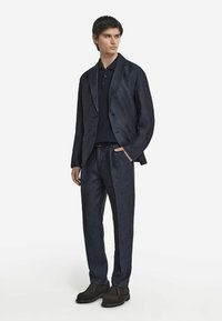 Mörk denimkostym med en figursydd kavaj och skräddarsydda byxor, kombinerad med en marinblå pikétröja. Bruna skor fulländar outfiten.