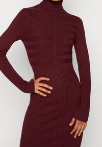 Abito a coste bordeaux con collo alto, caratterizzato da dettagli a righe verticali e una silhouette aderente. Maniche lunghe con polsini aderenti.