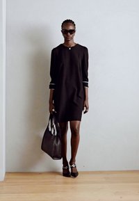 Black dress with three-quarter sleeves, white accents, paired with a black handbag and pointed-toe shoes. Model wearing sunglasses.