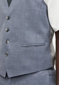 Ανδρικό ανοιχτό μπλε γιλέκο με υφή, με τέσσερα γκρι κουμπιά πάνω από ένα λευκό πουκάμισο, που δείχνει τσέπη στο στήθος και μέρος από τα ταιριαστά παντελόνια.