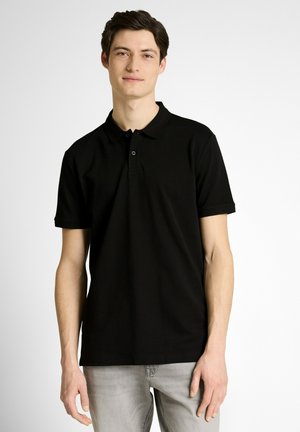 Junger Mann mit kurzen dunklen Haaren, der ein schwarzes kurzärmeliges Poloshirt und hellgraue Jeans trägt und vor einem schlichten weißen Hintergrund steht.