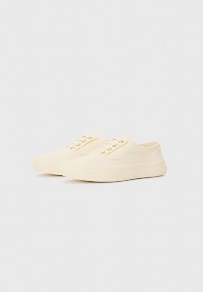 Sneakers in tela beige con punta arrotondata, suola piatta in gomma e occhielli dorati per i lacci. Design minimalista con texture liscia e branding discreto.
