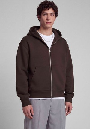 Giovane uomo che indossa una felpa con cappuccio marrone scuro con zip e tasche frontali, una maglietta bianca sotto, pantaloni grigio chiaro e una collana a catena d'argento.