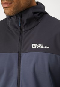 Navigasjonjakke med et design i mørk marineblå og lysere blå fargeblokker, glatt tekstur, høy krage og Jack Wolfskin-logo på brystet.