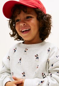 Χαμογελαστό παιδί με σγουρά μαλλιά, φοράει κόκκινο καπέλο και γκρι φούτερ με σχέδιο μικρών καλαθοσφαιριστών.