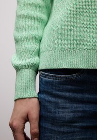 Main détendue le long du corps, portant un pull tricoté vert clair et un jean denim bleu foncé, sur un fond uni.