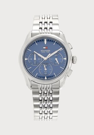 Montre-bracelet en métal argenté avec cadran bleu, trois sous-cadrans et logo "Hilfiger Dual Time" sous un petit drapeau rouge, blanc et bleu.
