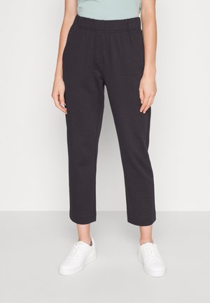 Person iført sorte elastiske bukser med kort ben og hvide sneakers, stående mod en ensfarvet lys baggrund.