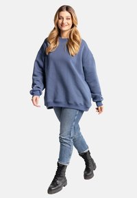 Niebieska, oversize bluza z okrągłym dekoltem i ściągaczami przy rękawach, zestawiona z jasnymi dżinsami i czarnymi butami wojskowymi. Gładka tekstura.