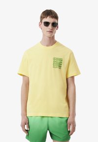 T-shirt en coton jaune avec un logo en texte vert répété sur la zone de la poche poitrine. Porté avec un short vert. Texture, coupe ample.
