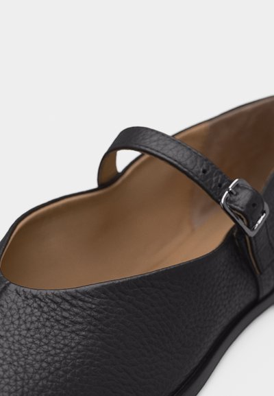 Chaussure plate Mary Jane en cuir noir texturé avec bride à boucle réglable et doublure intérieure beige sur fond blanc.