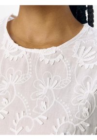 Haut blanc brodé présentant des motifs floraux et des coutures texturées, avec un col arrondi et des détails délicats sur le tissu.