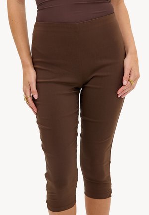 Femme portant un pantalon court marron foncé ajusté et un haut assorti, avec des bagues dorées sur les deux mains, debout sur un fond uni.