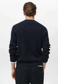 Mann in einem marineblauen, gerippten Strickpullover und dunklen Hosen, stehend mit den Händen in den Taschen, mit dem Rücken zur Kamera.