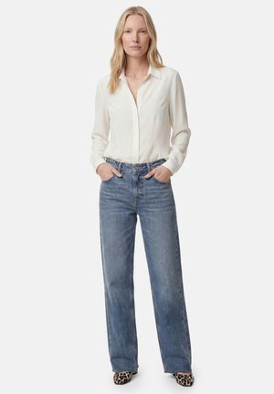 Blond kvinde står med hænderne i lommerne iført en hvid langærmet skjorte, blå jeans og leopardmønstrede sko på en ensfarvet baggrund.
