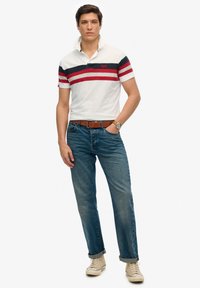 Hvid polo-shirt med røde og marineblå striber, kraven åben. Blå denim jeans og beige sneakers. Brun bælte og ur synligt. Casual pasform.