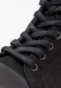Black leather shoe with textured finish, featuring thick black laces, metal eyelets, and a rounded toe design. Close-up view.