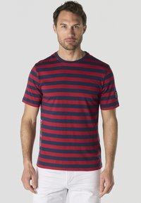 Homme portant une chemise à manches courtes avec des rayures horizontales rouges et bleu marine, et un pantalon blanc, debout devant un fond gris uni.