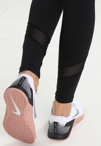 Valkoiset urheilukengät, joissa on mustia ja harmaita yksityiskohtia, teksturoitu päällinen, pinkki kumipohja, yhdistetty mustiin legginsseihin, joissa on verkkopaneeliaisia.