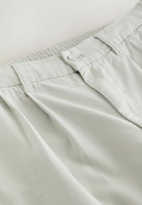 Gros plan sur la ceinture d'un pantalon gris clair avec fermeture par bouton, passants de ceinture et détail de ceinture élastique.