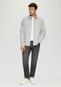 Hellgrau button-up Hemd mit langen Ärmeln und Kragen, kombiniert mit dunkelgrauen Jeans und weißen Sneakern vor einem schlichten Hintergrund.