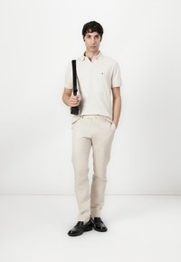 Lichtbeige poloshirt met korte mouwen, lichtbeige broek en zwarte schoenen. Draagt een zwarte schoudertas en staat tegen een witte achtergrond.