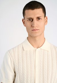 Ljusbeige kortärmad polo med strukturerade kabelstickade mönster och en ribbad krage med knappslå fram.