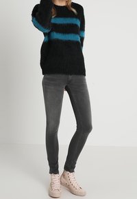 Persona vistiendo un suéter negro de felpa con rayas azules, jeans ajustados gris oscuro y zapatillas de caña alta rosa claro, de pie contra un fondo liso.