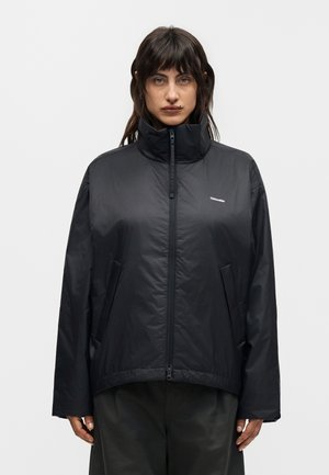 Άτομο που φοράει μαύρο μπουφάν με ψηλό γιακά, μπροστινό φερμουάρ και πλαϊνές τσέπες, στέκεται μπροστά σε απλό λευκό φόντο.