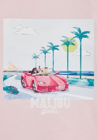 Rožinė marškinė su iliustracija, kurioje vaizduojami du žmonės raudoname kabrioletuose pakrantes kelio, apsupto palmėmis, saulėje ir užrašu "pagaminta MALIBU Barbie."