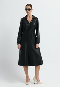 Žena v černých dlouhých rukávech z umělé kůže, s černými slunečními brýlemi, zlatými kruhovými náušnicemi a béžovými lodičkami na vysokém podpatku, stojící na bílém pozadí.